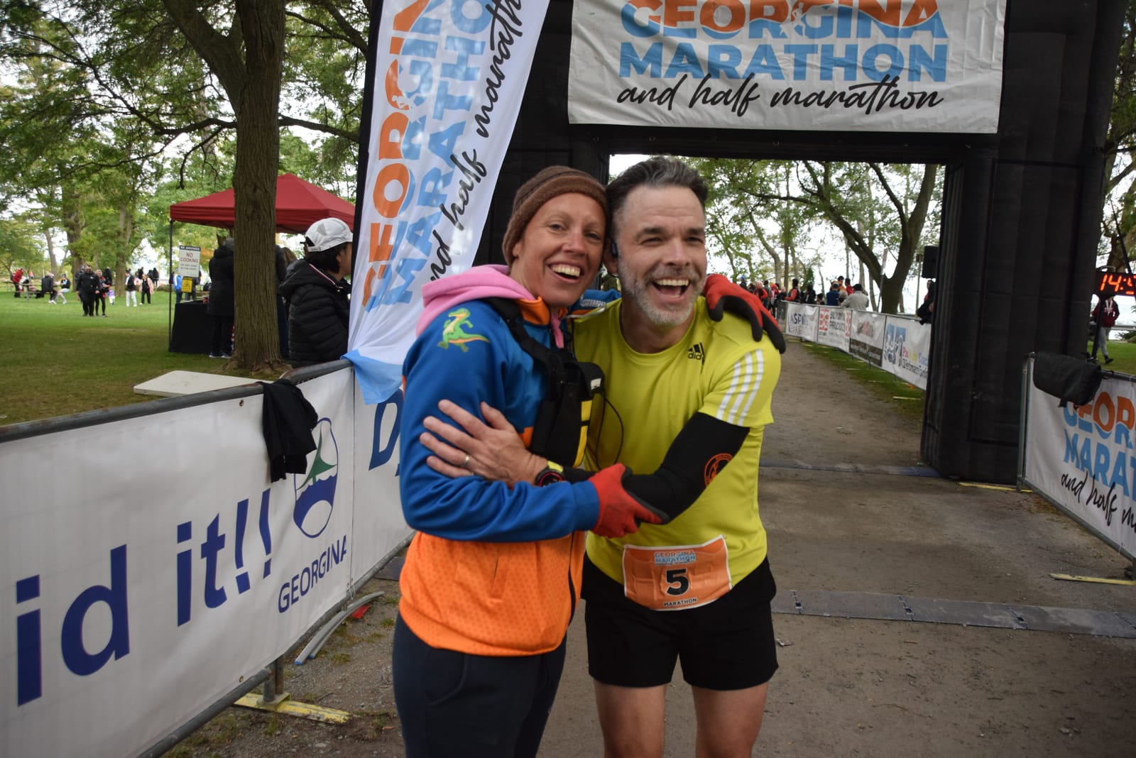 A runner with his coach smiling and hugging at the Georgina Marathon finish line. Bright sportswear, trees in the background, and marathon banners visible.