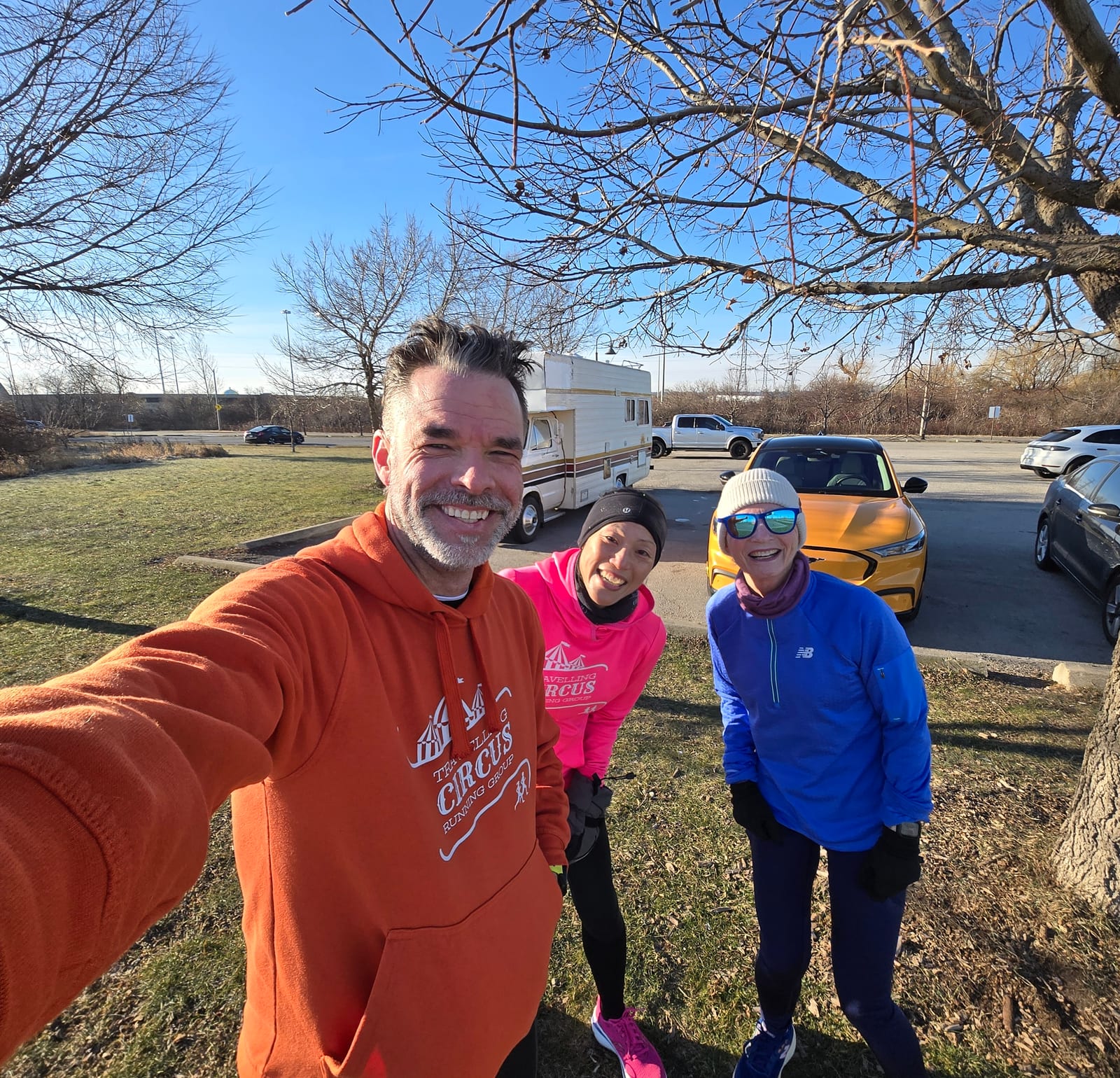 Three people in colorful hoodies smile for a selfie outdoors near parked cars. Bright blue sky, sparse trees, and grass in the background.