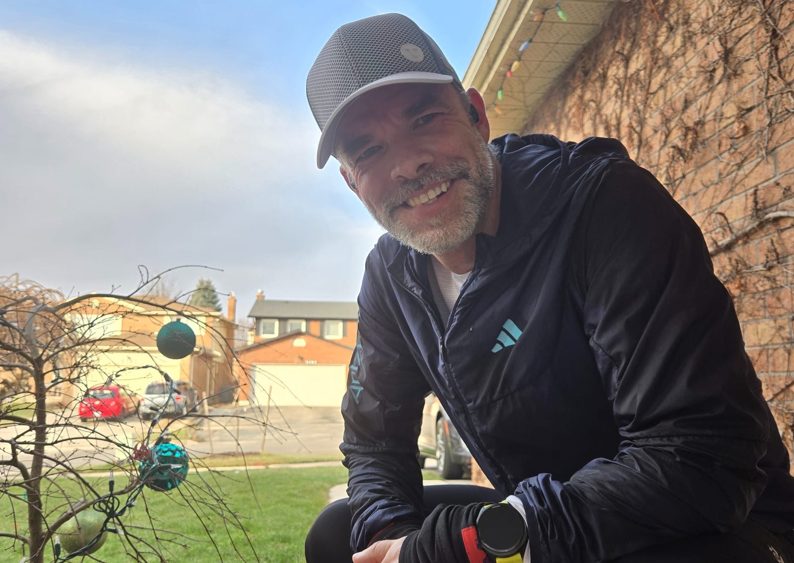 Man smiling in a cap and jacket, kneeling near a bare tree with blue ornaments. Background shows houses, cars, and a cloudy sky.