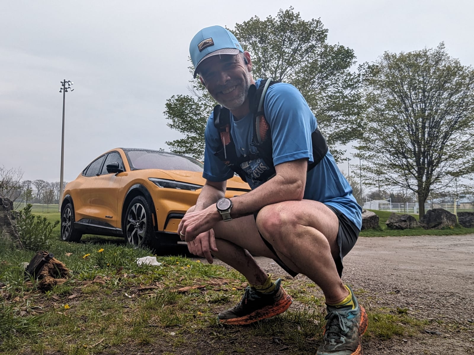 Man in blue squatting, smiling, wearing a cap and backpack. Yellow car behind him, with greenery and overcast sky in the background.