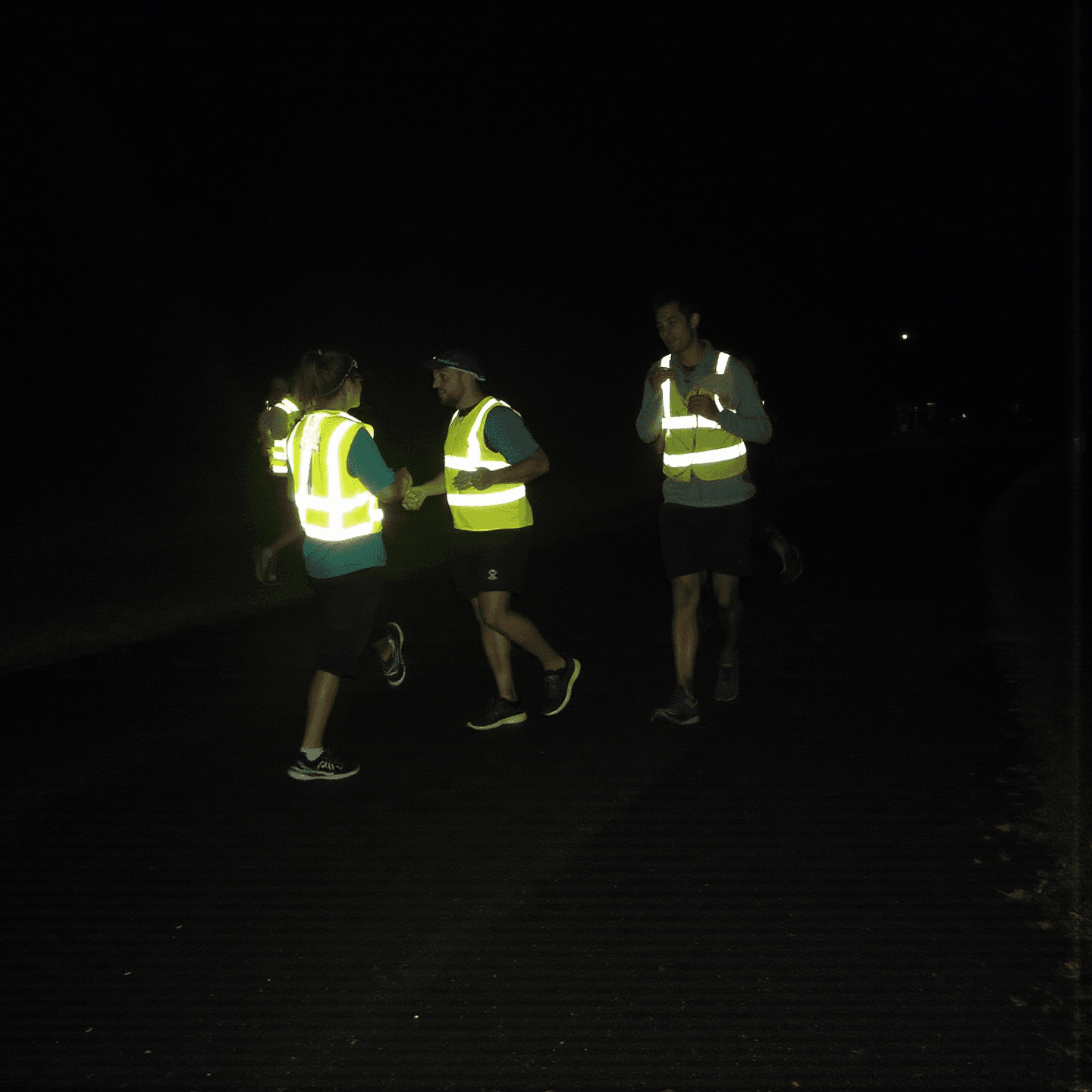 Runners in reflective vests jog on a dark path at night. The glowing vests stand out against the black background, creating a focused mood.