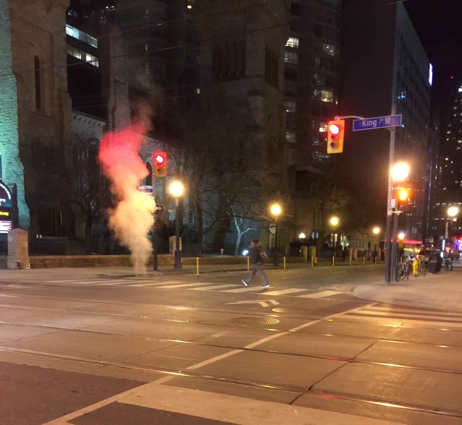Night city street; person jaywalking under a red light. Steam rises from a vent, illuminated by streetlights. Urban buildings in background.