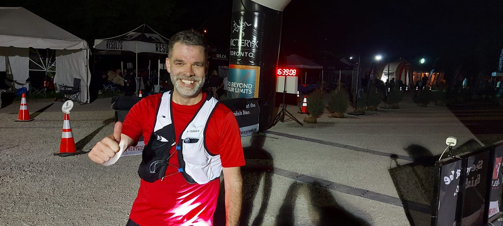 Doug Riches in red shirt and white vest smiling, giving thumbs up at nighttime race finish line. Timer shows 16:39:08. "Go Beyond Your Limits" sign.